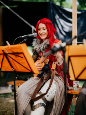 Ukulele mit Bina Bianca Eine lächelnde Musikerin mit rotem Haar, die ein Instrument spielt, während sie auf dem Elbenwald Festival auftritt. Sie trägt ein fantasievolles Kostüm mit Rüstungselementen und steht vor einem Mikrofon, umgeben von Notenblättern.