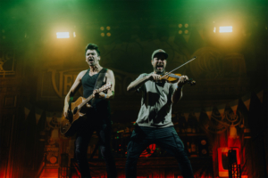 Versengold Zwei Musiker auf der Bühne des Elbenwald Festivals, einer spielt Gitarre und der andere Geige. Die Atmosphäre ist energiegeladen mit bunten Lichtern und einer begeisterten Menge im Hintergrund. Ein Highlight des nerdigen Musik- und Kulturereignisses.