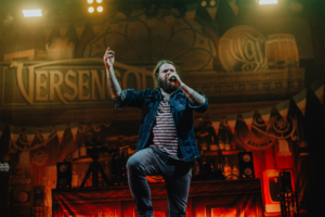 Versengold Ein Sänger auf der Bühne des Elbenwald Festivals, umgeben von bunten Lichtern und einer festlichen Kulisse. Er trägt ein gestreiftes Shirt und eine Jeansjacke, während er leidenschaftlich ins Mikrofon singt und das Publikum begeistert.