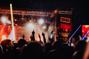 Versengold Bühne beim Elbenwald Festival mit einem leidenschaftlichen Musiker, umgeben von jubelnden Fans. Die Atmosphäre ist lebhaft, mit bunten Lichtern und erhobenen Händen, die die Freude und Energie des Events widerspiegeln.