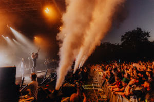 Versengold Live-Auftritt beim Elbenwald Festival mit begeistertem Publikum, Rauchmaschinen und stimmungsvollem Licht. Musiker mit Gitarre auf der Bühne, umgeben von jubelnden Fans, die die Atmosphäre eines einzigartigen Nerd-Events genießen.