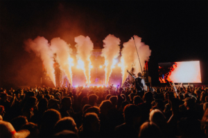 Versengold Bühne beim Elbenwald Festival mit bunten Lichtern und Rauchfontänen, umgeben von einer begeisterten Menge. Der Fokus liegt auf einem Performer, der das Publikum anfeuert, während die Atmosphäre festlich und energiegeladen ist.