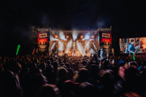 Versengold: Bunte Lichter und eine begeisterte Menge beim Elbenwald Festival, wo Musik und Nerd-Kultur aufeinandertreffen. Die Bühne ist mit Effekten beleuchtet, während Künstler auftreten und die Zuschauer mit leuchtenden Stäben feiern. Foto: Rainer Keuenhof