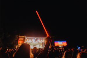 Versengold live Ein Festivalbesucher hebt ein leuchtendes rotes Lichtschwert in die Luft, während die Bühne im Hintergrund mit bunten Lichtern erleuchtet ist. Die Menge feiert bei Nacht auf dem Elbenwald Festival, einer einzigartigen Mischung aus Musik und Nerdkultur.