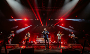 Versengold Live-Auftritt einer Band auf dem Elbenwald Festival, mit energiegeladenen Musikern auf der Bühne, umgeben von rotem Licht und einer lebhaften Festivalatmosphäre. Die Musiker spielen verschiedene Instrumente und begeistern das Publikum.