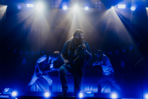 Versengold Ein Musiker steht im Rampenlicht des Elbenwald Festivals, umgeben von Bandmitgliedern. Die Bühne ist in blaues Licht getaucht, während er leidenschaftlich singt. Im Hintergrund sind Instrumente und die festliche Atmosphäre des Events erkennbar.