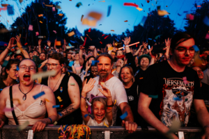 Versengold Buntes Festivalpublikum beim Elbenwald Festival, begeistert von der Musik, umgeben von Konfetti und jubelnden Fans. Die Atmosphäre ist lebhaft und voller Freude, während Menschen jeden Alters zusammen feiern und die besondere Stimmung genießen.