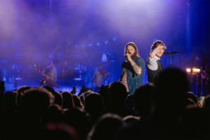 Versengold Zwei Sänger auf der Bühne beim Elbenwald Festival, umgeben von einer begeisterten Menge. Die Lichtshow und der Nebel schaffen eine energiegeladene Atmosphäre, während die Fans ihre Smartphones hochhalten, um den Moment festzuhalten.