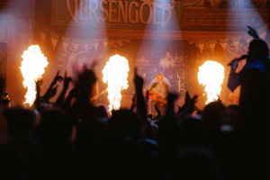 Versengold Bühne beim Elbenwald Festival mit Band und feurigen Effekten, umgeben von begeistertem Publikum. Die Atmosphäre ist lebhaft und energiegeladen, während die Musiker mit Instrumenten performen und die Menge mitreißen.