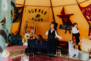 MMM Larp Eine Moderatorin steht auf der Bühne in einem bunten Zelt beim Elbenwald Festival, umgeben von Dekorationen und Bannern. Im Hintergrund sind verschiedene Requisiten und eine kleine Menschenmenge zu sehen, die aufmerksam zuschaut.