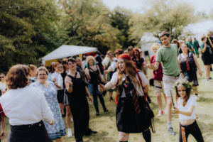 MMM Larp Teilnehmer des Elbenwald Festivals in Kostümen interagieren auf einer Wiese, während sie Zauberstäbe halten. Die Atmosphäre ist lebhaft und kreativ, mit einem bunten Publikum, das die Mischung aus Musik und Nerd-Kultur genießt.
