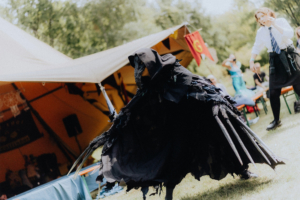 MMM Larp Ein kostümierter Teilnehmer in einem dramatischen schwarzen Outfit tanzt auf dem Elbenwald Festival. Im Hintergrund sind Zelte und andere Festivalbesucher zu sehen, die die lebendige Atmosphäre der Veranstaltung genießen.