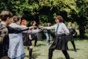 MMM Larp Eine Gruppe von Teilnehmern in Kostümen führt einen Zauberduell-Workshop auf dem Elbenwald Festival durch, wobei sie mit Zauberstäben agieren. Die Atmosphäre ist lebhaft und kreativ, umgeben von grünem Hintergrund und anderen Festivalbesuchern.