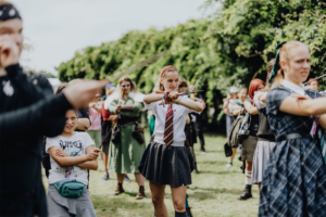 MMM Larp Eine Gruppe von Festivalbesuchern in Kostümen übt eine Choreografie auf dem Elbenwald Festival. Die Teilnehmer zeigen Kreativität und Begeisterung, während sie eine Mischung aus Musik und Fantasie erleben. Grüne Bäume im Hintergrund verleihen der Szene eine lebendige Atmosphäre.