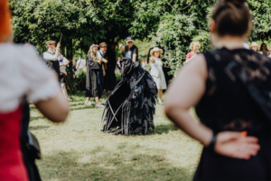 MMM Larp Ein Teilnehmer in einem aufwendigen Kostüm steht im Mittelpunkt des Elbenwald Festivals, während eine Gruppe von Zuschauern in fantasievoller Kleidung ihn beobachtet. Die Atmosphäre ist lebhaft, mit Bäumen im Hintergrund und Sonnenlicht, das auf die Szene fällt.