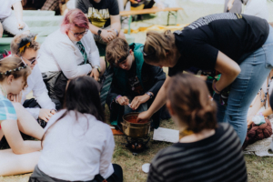 Eine Gruppe von Festivalbesuchern beim Elbenwald Festival, die um einen kleinen Kochtopf sitzen und gemeinsam eine kreative Aktivität durchführen. Die Teilnehmer tragen fantasievolle Kostüme und genießen die gemeinschaftliche Atmosphäre.