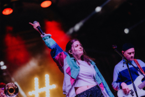 #zweiraumsilke Auftritt einer Sängerin mit Mikrofon auf der Bühne des Elbenwald Festivals, umgeben von bunten Lichtern. Im Hintergrund ist ein Musiker am Bass zu sehen. Die Atmosphäre ist lebhaft und voller Energie, perfekt für das Publikum.