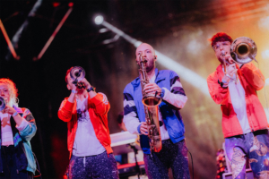 #zweiraumsilke Vier Musiker in bunten Outfits spielen Trompeten und ein Saxophon auf der Bühne des Elbenwald Festivals. Die energiegeladene Atmosphäre wird durch farbenfrohe Lichter und eine begeisterte Menge verstärkt, die die einzigartige Mischung aus Musik und Nerd-Kultur genießt.