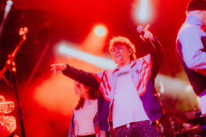 #zweiraumsilke Ein Sänger auf der Bühne des Elbenwald Festivals, umgeben von rotem Licht und einer energiegeladenen Atmosphäre, während er das Publikum begeistert und seine Performance genießt. Im Hintergrund sind weitere Musiker und Instrumente sichtbar.