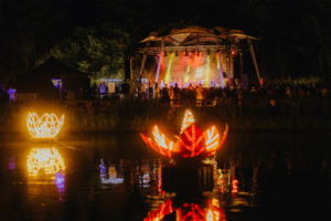 #zweiraumsilke Bunt beleuchtete Bühnenatmosphäre beim Elbenwald Festival, mit Live-Musik und neugierigen Besuchern. Im Vordergrund reflektieren leuchtende Wasserpflanzen das Festivalgeschehen, während im Hintergrund die Bühne mit Künstlern zu sehen ist.