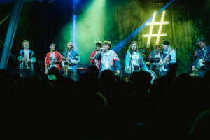 #zweiraumsilke Bühne beim Elbenwald Festival mit einer Band, die live auftritt. Musiker in bunten Jacken spielen verschiedene Instrumente, während das Publikum begeistert zuschaut. Bunte Lichter und eine grüne Kulisse schaffen eine festliche Atmosphäre.