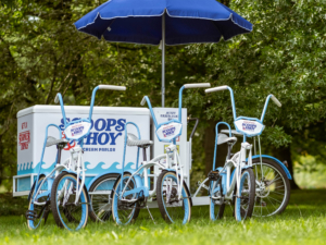 Drei Fahrräder stehen vor einem Eiswagen mit dem Logo "Scoops Ahoy" aus der Serie "Stranger Things". Der Wagen ist mit einem blauen Sonnenschirm ausgestattet und befindet sich auf einer grünen Wiese, umgeben von Bäumen. Ideal für das Elbenwald Festival.