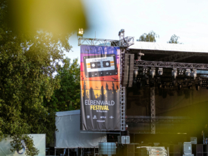 Banner des Elbenwald Festivals mit einem nostalgischen Kassettendesign, das die Verbindung von Musik und Fantasie symbolisiert, vor einer Bühne mit Lichttechnik und Bäumen im Hintergrund. Perfekte Kulisse für ein einzigartiges Nerd-Event.