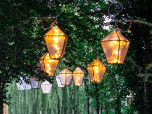 Schöne, beleuchtete Laternen hängen an einem grünen Baumgang beim Elbenwald Festival. Die warmen Lichter schaffen eine einladende Atmosphäre und verleihen dem Festival eine magische Note, die Musik- und Nerdkultur vereint.