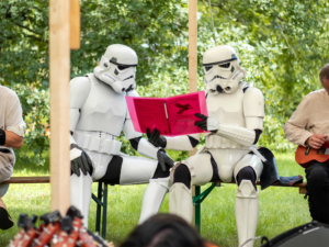 Zwei Stormtrooper in Kostümen sitzen auf einer Bank und studieren ein pinkes Liederbuch beim Elbenwald Festival, umgeben von grüner Natur und einem musikalischen Ambiente. Die Szene verbindet Cosplay mit musikalischen Darbietungen.