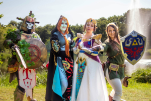 Cosplay-Gruppe auf dem Elbenwald Festival, bestehend aus vier Charakteren: einem kriegerischen Ritter, einer mysteriösen Figur, einer Prinzessin und einem Helden mit Schild. Die Szene spielt im Freien vor einem Teich, umgeben von Natur und Sonnenschein.