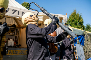 Auftritt von Künstlern in fantasievollen Kostümen beim Elbenwald Festival. Die Musiker tragen Masken und spielen auf ungewöhnlichen Instrumenten, während das Publikum die kreative Atmosphäre des Festivals genießt.
