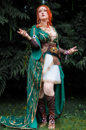 Kostümierte Teilnehmerin beim Elbenwald Festival, gekleidet in ein grünes, kunstvoll verziertes Outfit mit goldenen Akzenten und Stiefeln, posiert vor einer grünen Kulisse. Die Verbindung von Musik und Fantasie wird durch die lebendige Darstellung unterstrichen.