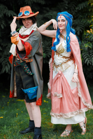 Zwei Teilnehmer in fantasievollen Kostümen beim Elbenwald Festival, einem Mix aus Musikfestival und Convention. Der Charakter links trägt einen Hut und eine abenteuerliche Kleidung, während die Teilnehmerin rechts in einem eleganten, historischen Outfit mit blauen Haaren posiert.