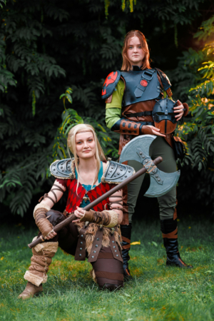 Zwei Cosplayer in fantasievollen Kostümen posieren auf dem Elbenwald Festival. Die erste Person kniet mit einem großen Axt-Requisit, während die zweite Person im Hintergrund steht und einen Helm hält. Die grüne Umgebung ergänzt die kreative Atmosphäre des Festivals.