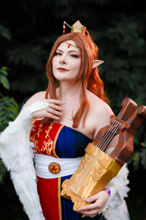 Cosplayerin in detailreicher Kostümierung mit einem Musikinstrument beim Elbenwald Festival, umgeben von grüner Natur. Die Figur verkörpert einen Charakter aus der Fantasiewelt, was den nerdigen und kreativen Geist des Festivals widerspiegelt.