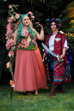 Zwei Frauen in fantasievollen Kostümen stehen auf einer Wiese. Die erste trägt ein blütenverziertes Outfit mit einem Stab, während die zweite eine Kapuze und ein Schwert hält. Die Farben und Details der Kostüme reflektieren die kreative Atmosphäre des Elbenwald Festivals.