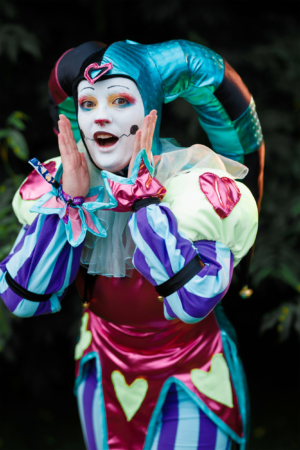 Bunt kostümierte Person mit Clown-Makeup und fröhlicher Pose auf dem Elbenwald Festival, umgeben von grüner Natur. Die Veranstaltung vereint Musik und Nerd-Kultur und zieht zahlreiche Besucher in fantasievollen Outfits an.