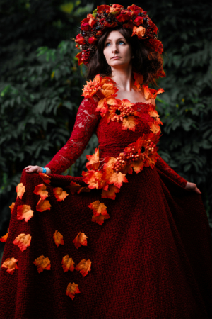 Eine Frau in einem roten Kleid, geschmückt mit blühenden Herbstblättern und einer Blumenkrone, posiert im Freien. Die lebendigen Farben und das kreative Outfit spiegeln die fantasievolle Atmosphäre des Elbenwald Festivals wider.
