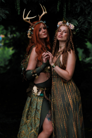 Zwei Frauen in fantasievoller Naturkleidung posieren unter grünen Blättern. Die eine trägt Geweih und Blumenkranz, die andere hat geflochtenes Haar und ein elegantes Kleid. Ihr Outfit spiegelt die kreative Atmosphäre des Elbenwald Festivals wider.