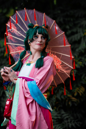 Frau in traditioneller Kleidung mit grünem Haar und einem dekorativen Regenschirm, posiert vor einer grünen Kulisse beim Elbenwald Festival, das Musik und Nerdkultur vereint. Die Farben ihres Outfits und der Regenschirm schaffen eine lebendige Atmosphäre.