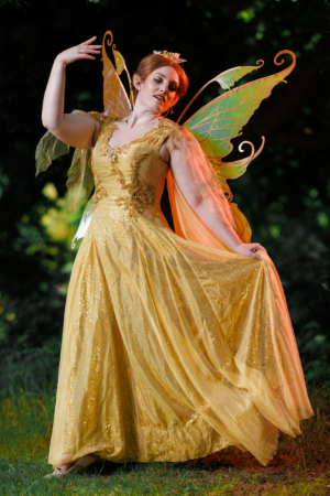 Eine Frau in einem glitzernden, goldenen Kleid mit transparenten, bunten Flügeln posiert im Freien. Die lebendige Szenerie und ihr kreatives Kostüm spiegeln die fantasievolle Atmosphäre des Elbenwald Festivals wider.