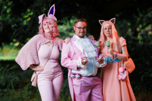 Drei Personen in bunten Kostümen beim Elbenwald Festival, umgeben von Natur. Die Teilnehmer tragen pinke Outfits und Accessoires, darunter Plüschfiguren und Mikrofone, und zeigen eine lebendige, kreative Atmosphäre des Festivals.