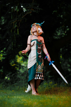 Cosplayer auf dem Elbenwald Festival in einem aufwendigen Kostüm mit grünen und weißen Elementen, verziert mit Holzperlen und einem Schwert in der Hand. Die Szene zeigt eine lebendige Verbindung von Fantasy und Natur im Hintergrund.