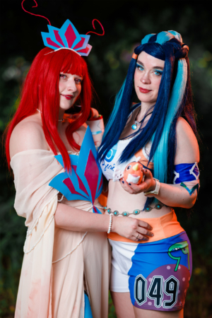 Zwei Cosplayerinnen in farbenfrohen Kostümen posieren beim Elbenwald Festival. Die eine trägt ein elegantes Outfit mit rotem Haar und einer dekorativen Krone, während die andere in einem sportlichen Look mit blauen Haaren und einem Spielzeug in der Hand strahlt.