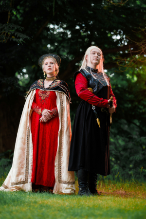 Zwei verkleidete Teilnehmer des Elbenwald Festivals stehen im Freien. Die Frau trägt ein rotes Kleid mit goldenen Verzierungen und einer eleganten Kapuze, während der Mann in einer dunklen Rüstung mit Schwert zu sehen ist. Die Szenerie vermittelt eine fantasievolle Atmosphäre.