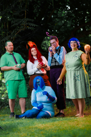 Fünf Personen in bunten Kostümen posieren auf einer Wiese beim Elbenwald Festival. Die Charaktere haben leuchtende Haare und halten verschiedene farbige Kugeln. Die Szene strahlt eine fröhliche, kreative Atmosphäre aus, die das Festivalfeeling widerspiegelt.