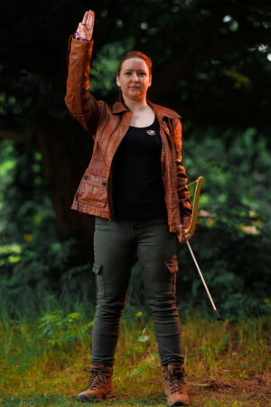 Eine Person in brauner Lederjacke und schwarzem T-Shirt hebt die Hand in einer charakteristischen Geste. Sie trägt grüne Cargo-Hosen und Wanderschuhe, steht auf einer Wiese mit Bäumen im Hintergrund. Die Szene ist lebhaft und spiegelt die kreative Atmosphäre des Elbenwald Festivals wider.