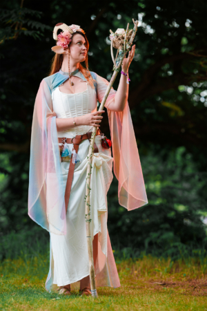 Eine Frau in einem fantasievollen Kostüm mit einem Stab steht auf einer Wiese beim Elbenwald Festival. Ihr Outfit besteht aus einem weißen Kleid und einem schimmernden Umhang, ergänzt durch Blumen und Accessoires, die eine magische Atmosphäre schaffen.