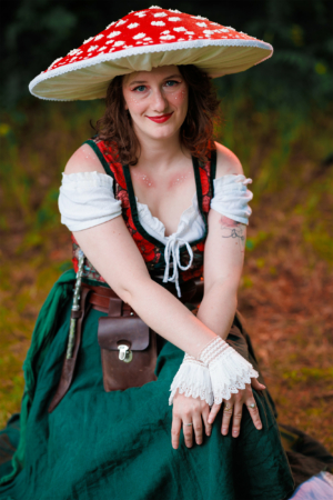 Eine junge Frau sitzt im Freien und trägt ein fantasievolles Kostüm mit einem großen, roten Pilzhut und einem grünen Rock. Ihre Hände sind mit zarten, weißen Spitzenärmeln geschmückt. Der Hintergrund zeigt eine natürliche, grüne Umgebung des Elbenwald Festivals.