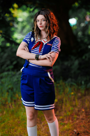 Ein cosplaying Fan auf dem Elbenwald Festival trägt ein blaues Outfit mit Streifen und roter Krawatte. Sie posiert selbstbewusst im Freien, umgeben von natürlichem Licht und grüner Vegetation, was die kreative Atmosphäre des Festivals unterstreicht.